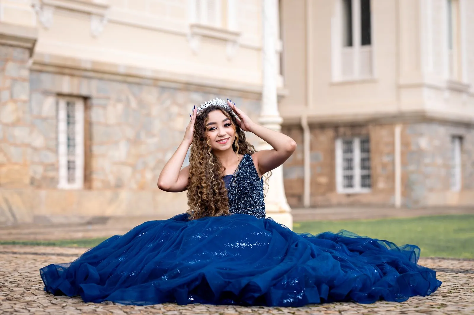 A woman in a stunning blue gown and tiara sits elegantly outside a historic building.